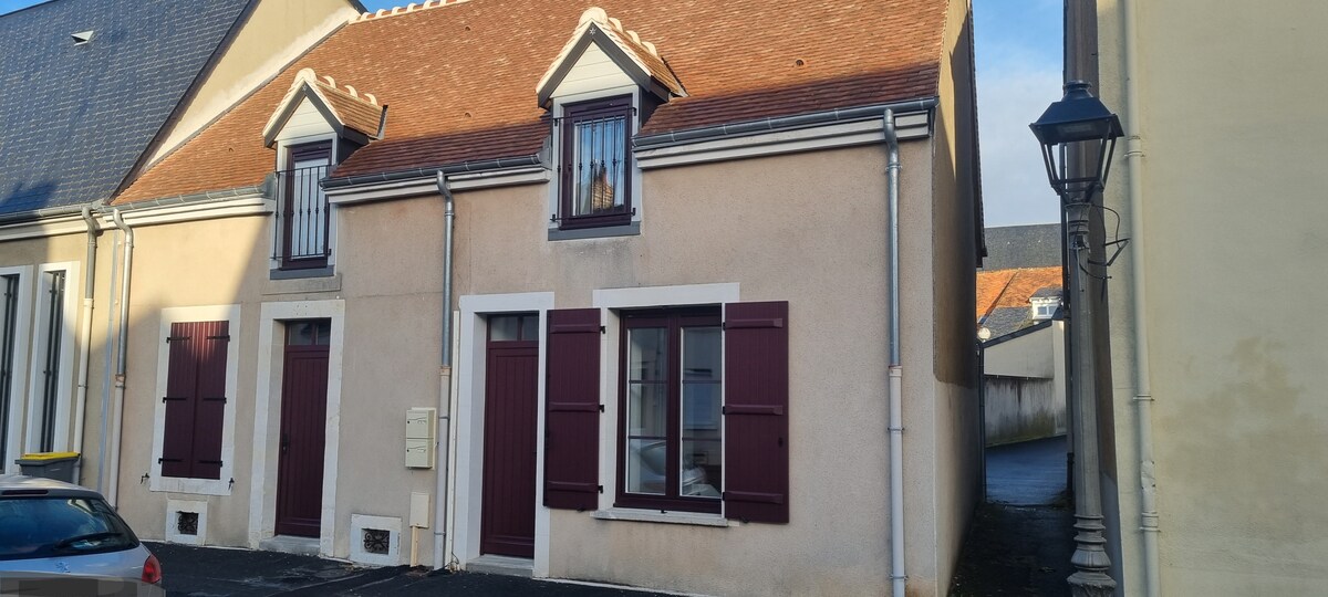 The exterior of a charming cottage showcases a light-colored facade with white trim and burgundy shutters. Two symmetrical windows adorn the front, each flanked by decorative shutters, while a brown-tiled roof adds a traditional touch. A street lamp stands nearby, complementing the setting.