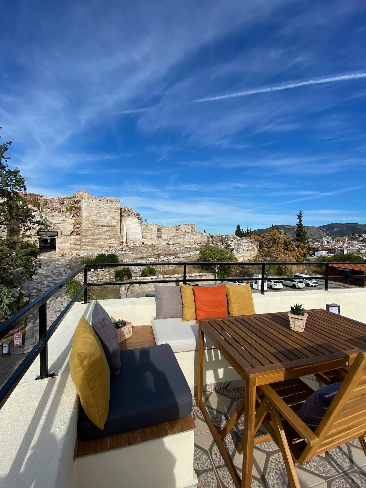 A terrace is displayed featuring a wooden dining table surrounded by two chairs and a cozy seating area with colorful cushions. A scenic view of ancient ruins and vibrant trees is visible in the background under a clear blue sky.