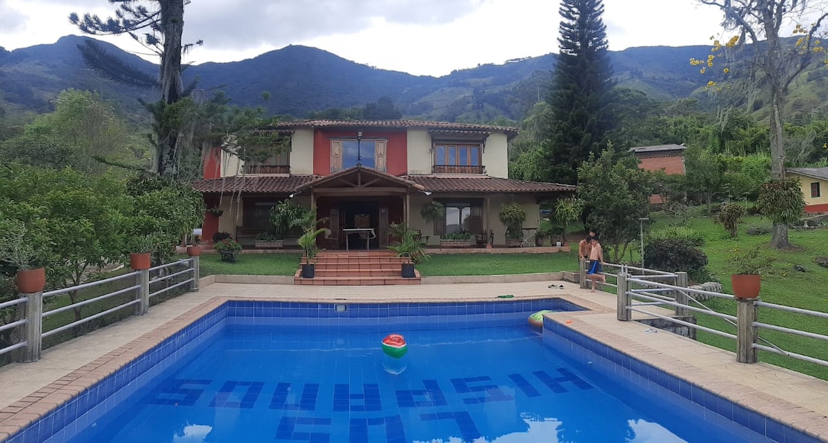 A tranquil outdoor scene features a swimming pool surrounded by well-maintained gardens. The two-story house is visible in the background, with a tiled roof and vibrant window shutters. Lush mountains rise in the distance, enhancing the serene setting.