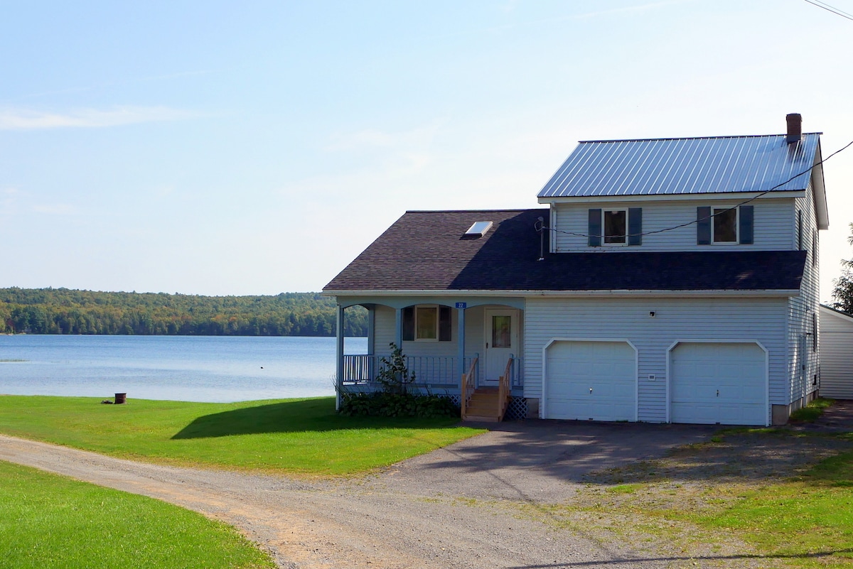 A two-story cabin is seen at the edge of Deering Lake, featuring a light-colored exterior and a metal roof. The front porch is accessible by a set of steps. Expansive lawns lead to the serene waterfront, surrounded by trees lining the shoreline.