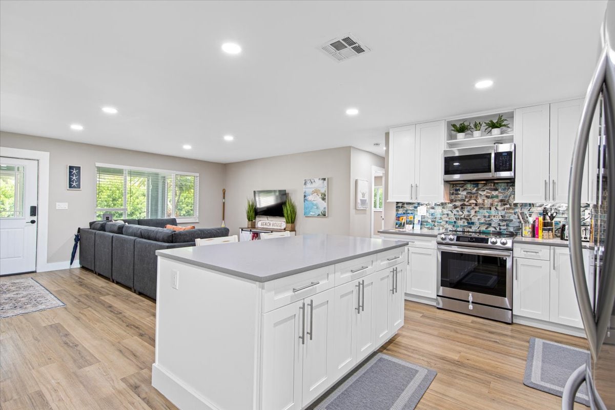 An open kitchen and living area are showcased, featuring modern appliances and a large island with a light countertop. Natural light fills the space through several windows, while comfortable seating is arranged in the living area, designed for relaxation and socializing.