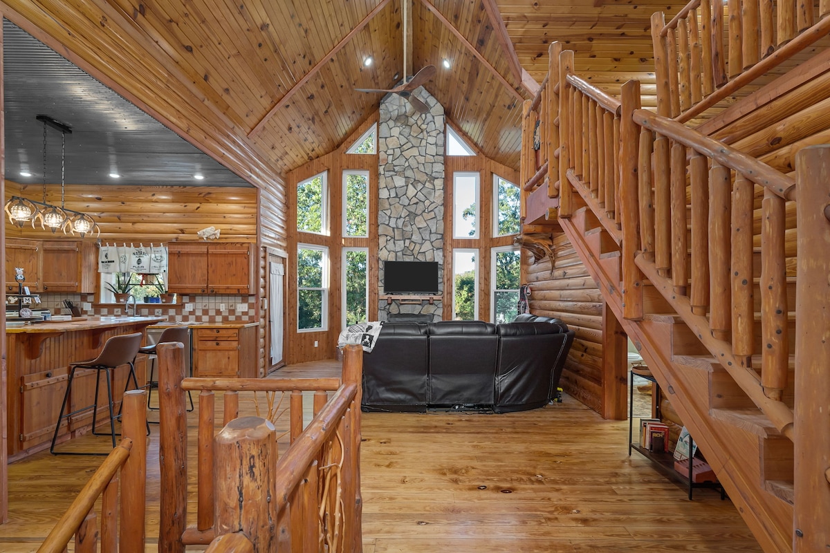 The interior showcases a spacious living area with high ceilings and log cabin aesthetics. Large windows allow natural light to flood in, highlighting the stone fireplace and wood finishes. A staircase leads to the upper level, while the kitchen and dining area are visible in the background.