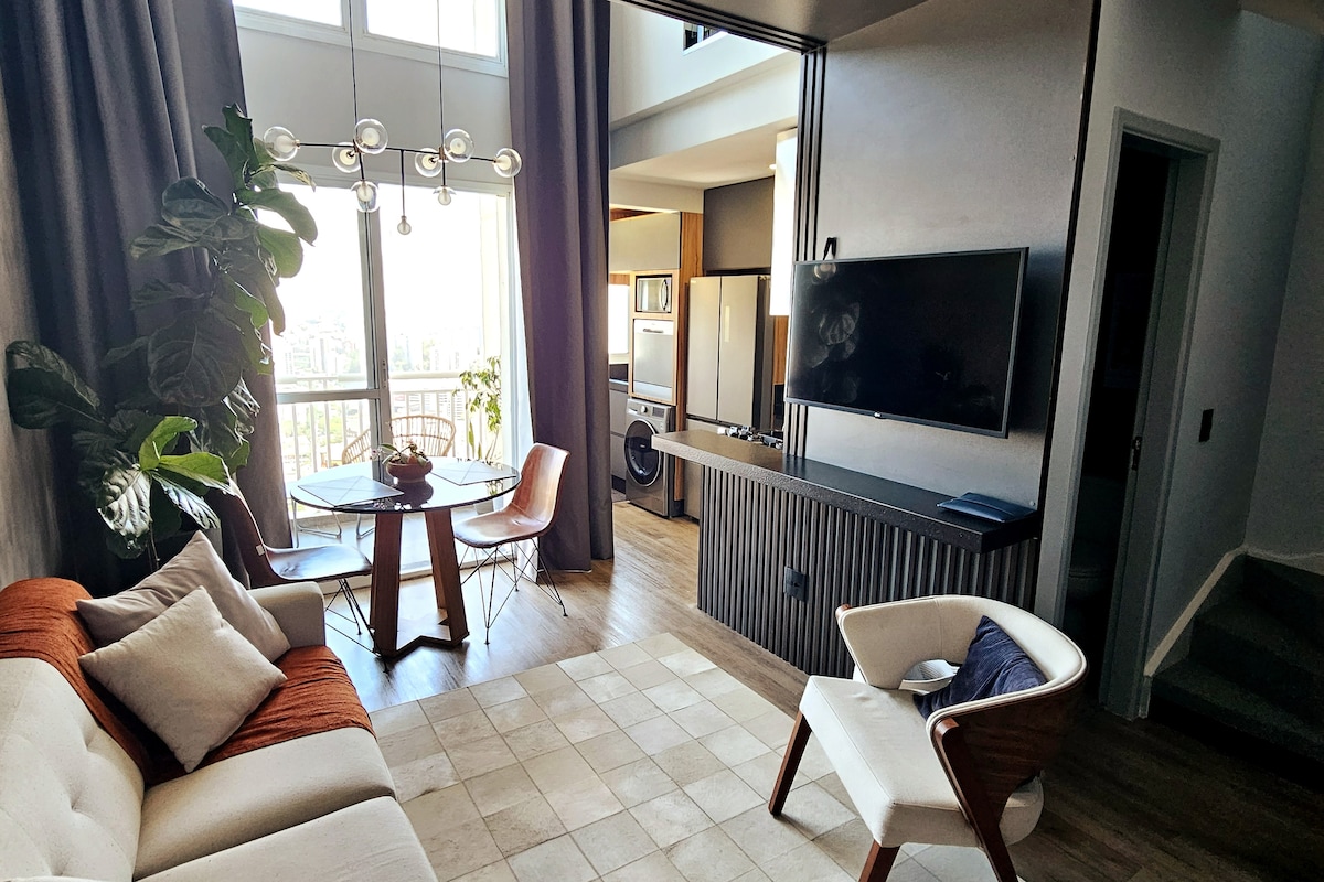 A modern living area features a comfortable sofa, a round dining table with stylish chairs, and a large television mounted on the wall. Tall windows allow natural light to illuminate the space, while indoor plants add a touch of greenery.