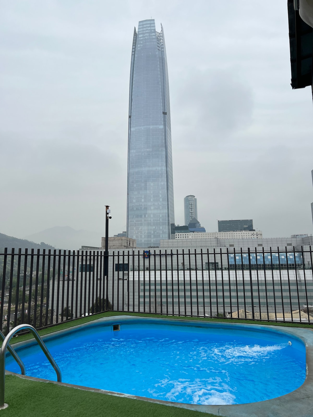 A rooftop pool is visible, featuring clear blue water that contrasts with the surrounding gray sky. The backdrop showcases a prominent skyscraper, its sleek glass facade reflecting the urban environment, enclosed by a black railing adding an element of safety.