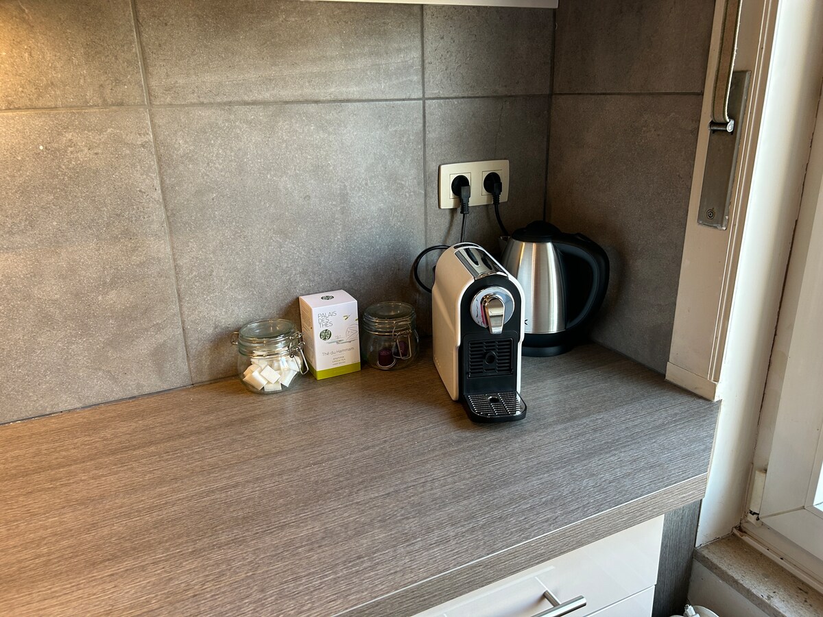 A modern kitchen counter is displayed with a coffee machine, a kettle, and jars containing sugar cubes and other small items. A small box of tea bags is visible, set against a backdrop of textured gray tiles and a window providing natural light.