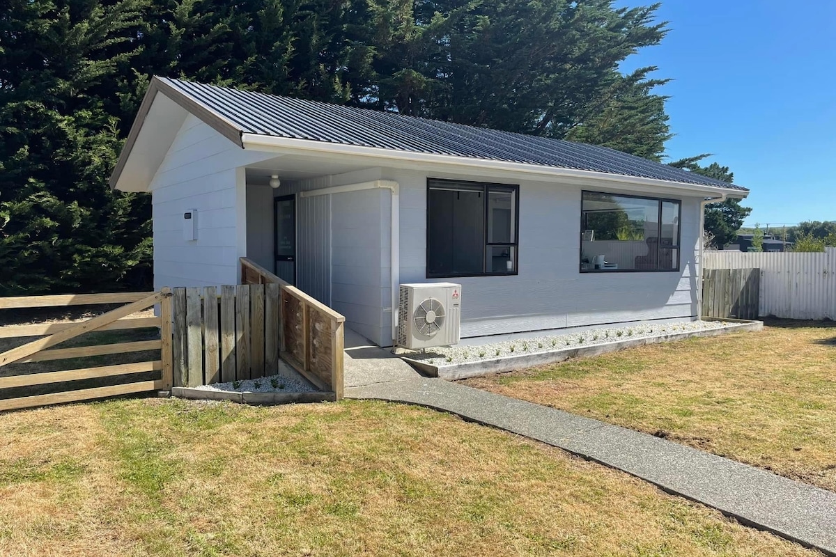 A single-story unit is set on a fully fenced section, featuring a combination of light-colored siding and a dark roof. A pathway leads to the entrance, surrounded by grass and bordered by wooden fencing. Large windows provide natural light to the interior.