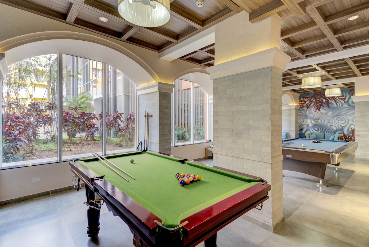 A billiards room is presented, featuring a green felt pool table with cues and balls prepared for play. Natural light filters in through large windows, illuminating the space adorned with stylish ceiling fixtures. A separate pool table is positioned in the background, enhancing the recreational atmosphere.