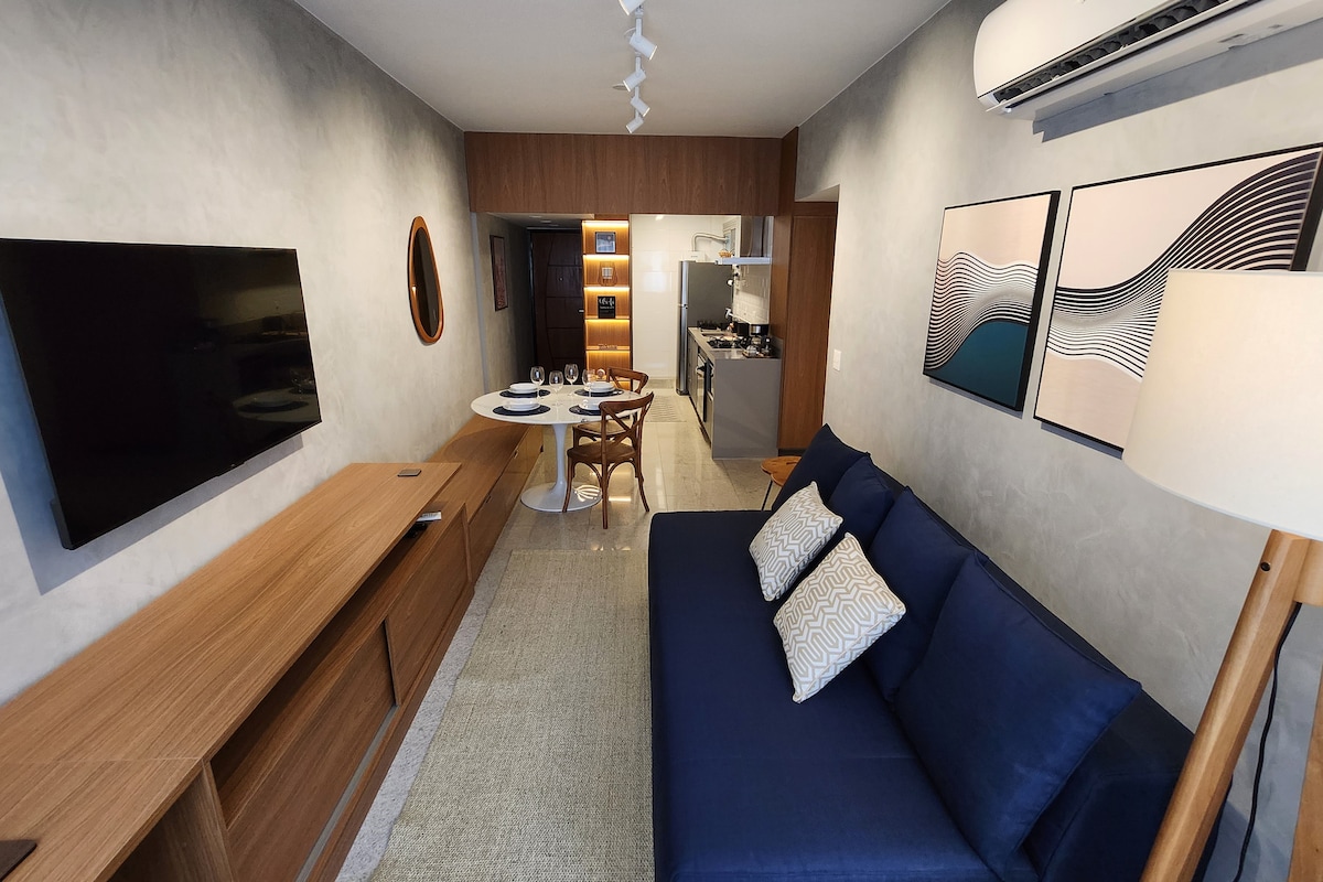 A modern living space features a navy blue sofa with decorative cushions, a round dining table set for four, and a large wall-mounted television. Neutral tones and textured walls create a calming environment, with artwork enhancing the decor. A well-equipped kitchen is visible in the background.