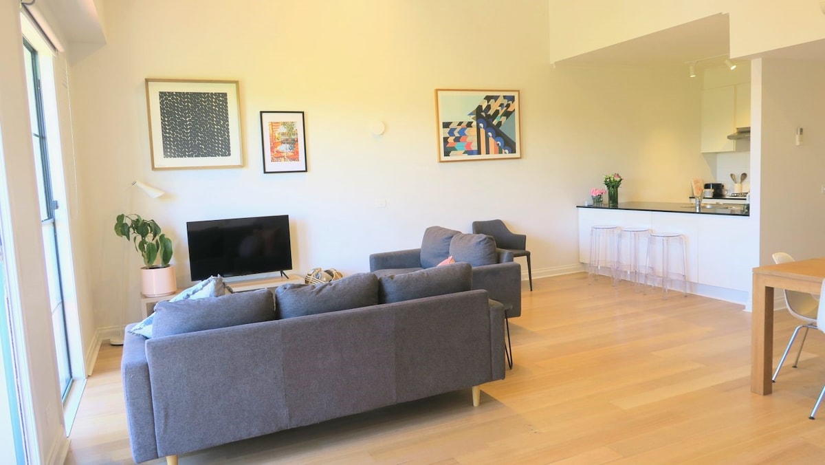 A spacious living area is filled with natural light, featuring two gray sofas and a television on a media unit. The open plan layout connects to the kitchen and dining area, showcasing a wooden dining table and clear acrylic chairs. Artwork adorns the walls.