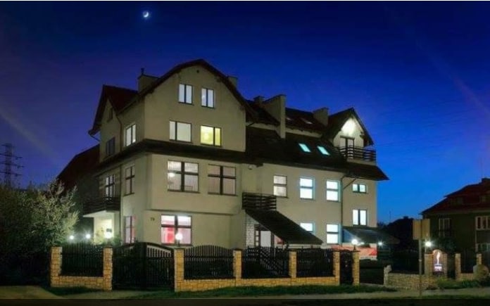 The exterior of a three-story building is illuminated against a twilight sky, showcasing multiple windows that create a warm glow. A surrounding fence adds privacy, and the structure features a combination of balconies and unique architectural details.