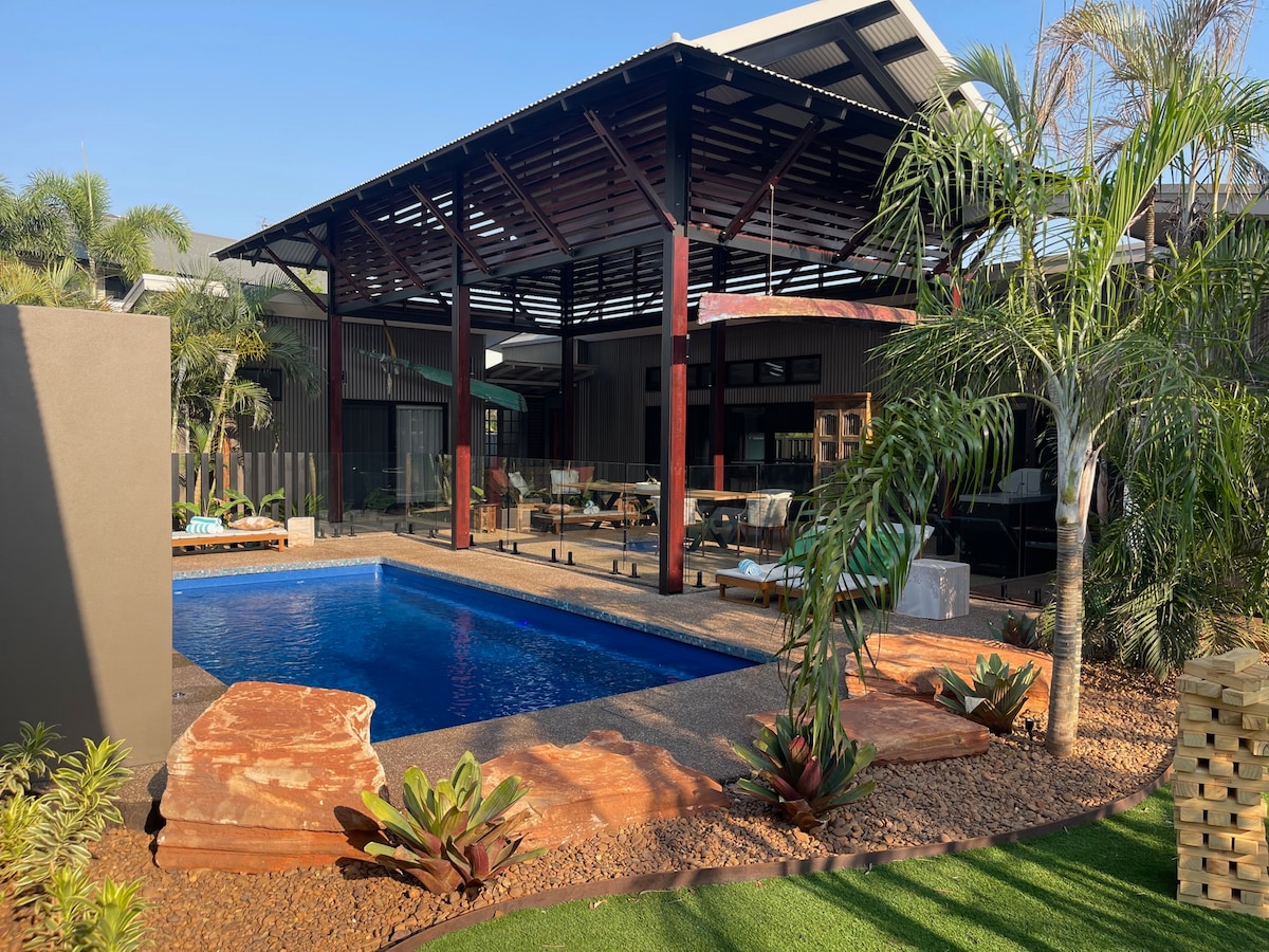 A tranquil outdoor area features a sparkling pool surrounded by lush greenery and decorative stone landscaping. A covered seating area is visible in the background, providing shade. The space is designed for relaxation, showcasing a blend of natural materials and modern architecture.