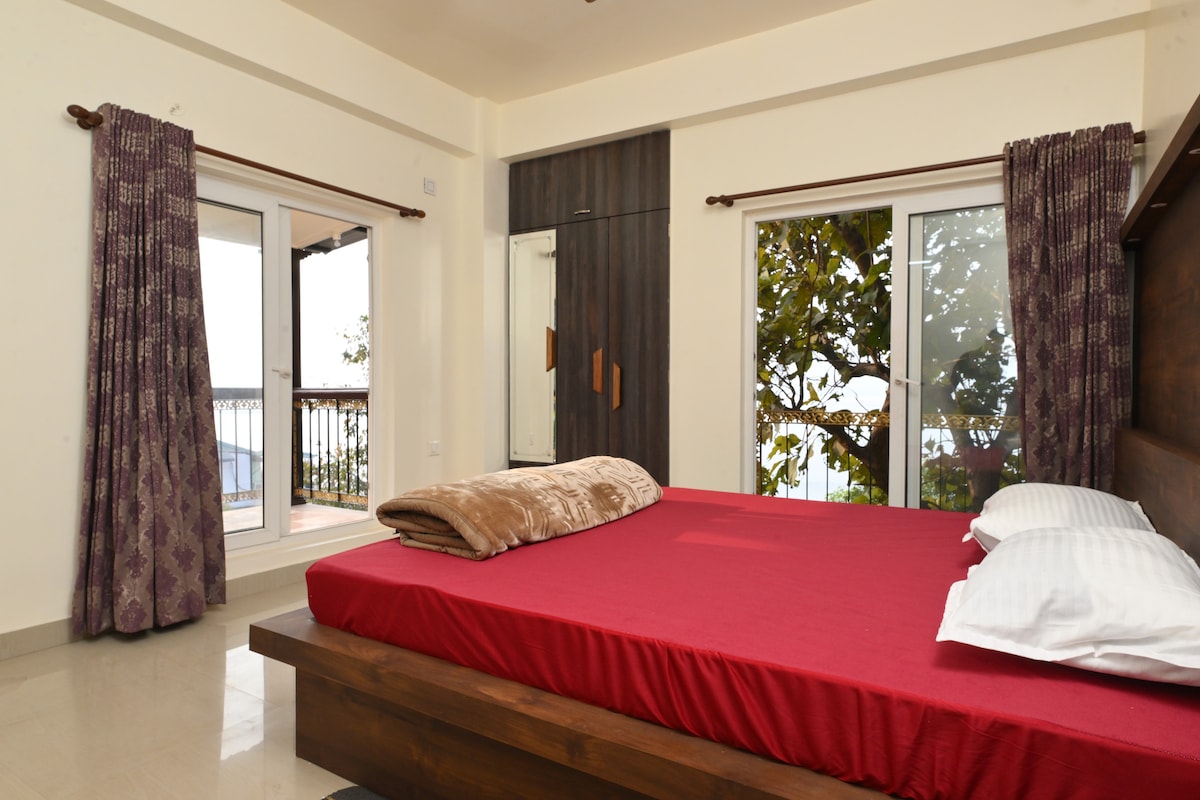 A spacious bedroom features a large wooden bed dressed with a maroon blanket. Light flows through two windows, showcasing views of the outside greenery. Dark curtains frame the windows, and a wooden wardrobe is visible along the wall.
