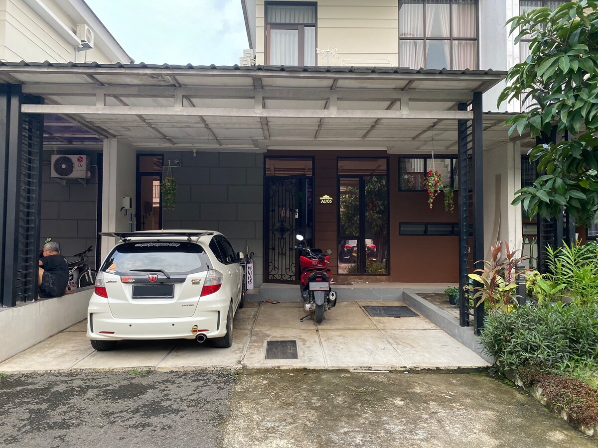 A covered parking area is visible, accommodating a white car and a motorcycle. The space is bordered by plants, providing a natural touch. The entrance to the house can be seen in the background, featuring a decorative gate and a welcoming ambiance.