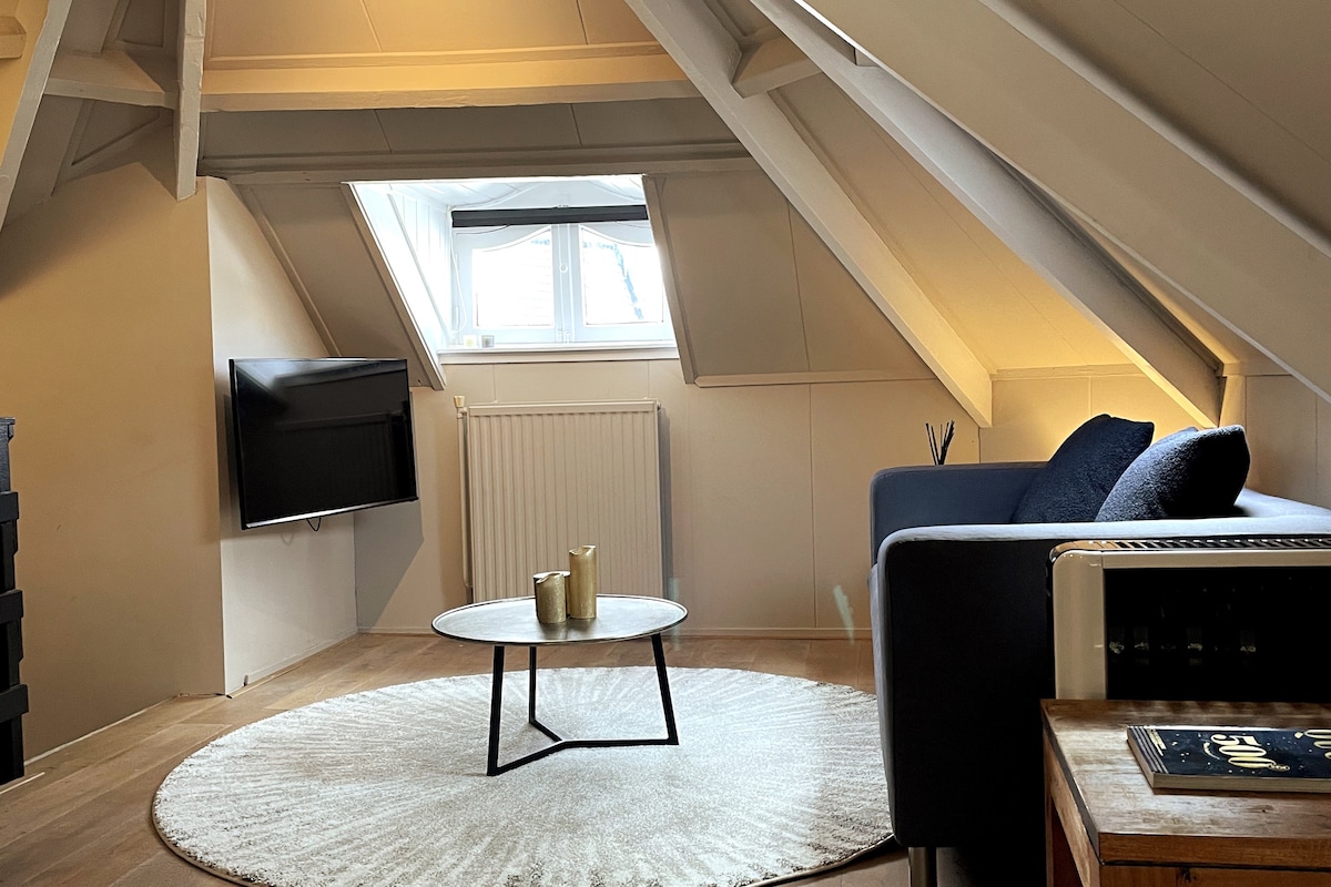 A cozy attic living area features sloped ceilings with wooden beams. A comfortable couch is positioned beside a round coffee table on a soft area rug. A flat-screen television is mounted on the wall, and light enters through a small window with a dark curtain.