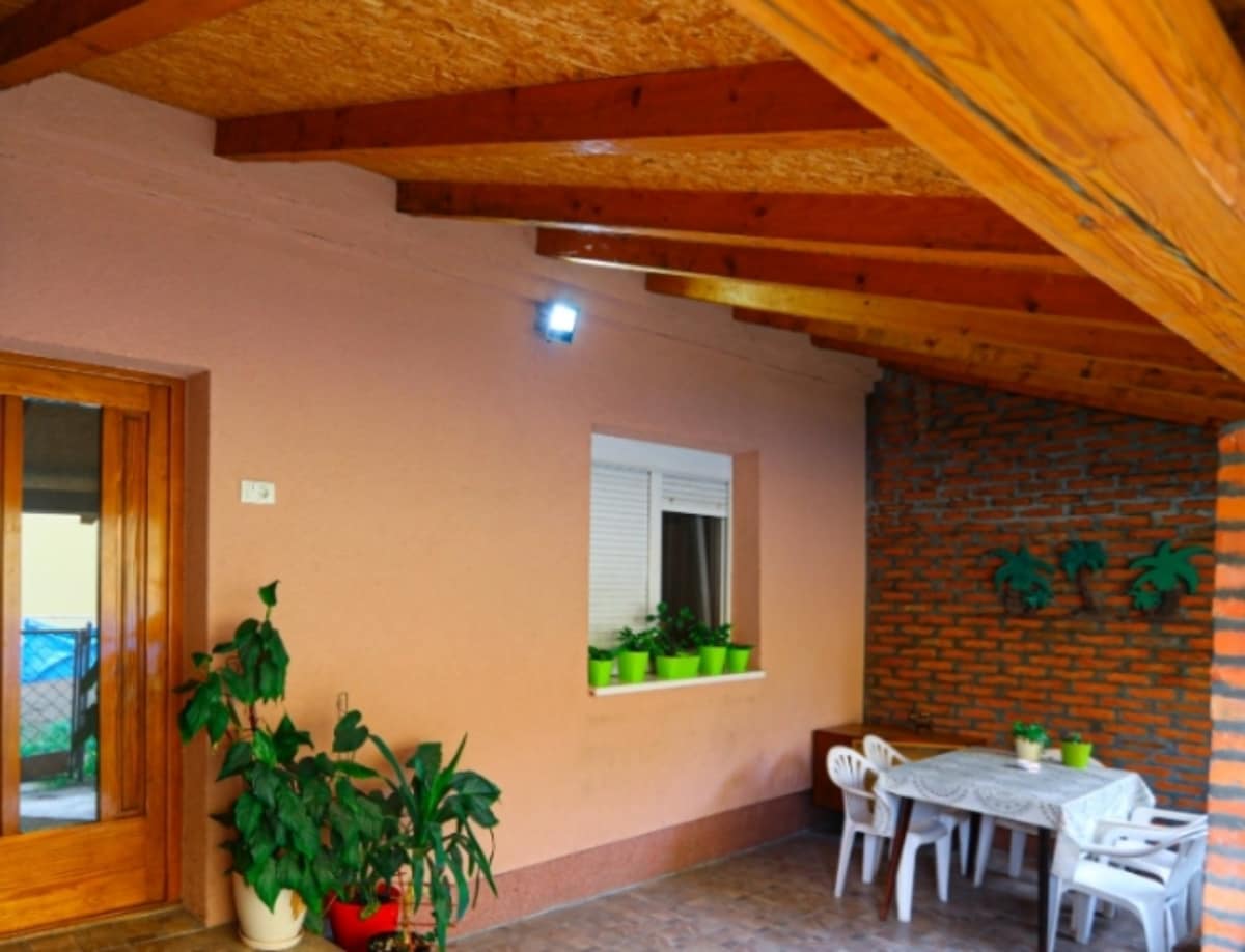 A covered patio space is displayed with wooden beams overhead. A small table set is visible, accompanied by white plastic chairs. Potted plants line the windowsill, while a brick wall adds texture to the area, offering a sense of serenity.