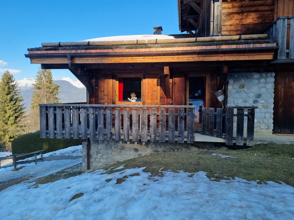 A wooden balcony with a rustic railing overlooks a snowy landscape. The chalet's facade features traditional wooden elements and stonework, with hints of green grass emerging near the snow. Mountains are visible in the distance under a bright blue sky.