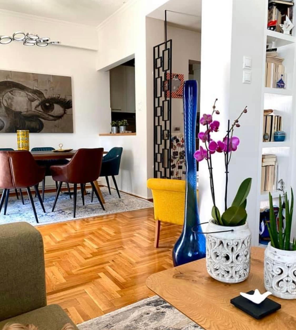 An inviting living area features a mix of modern furniture and vibrant decor. A hardwood floor is complemented by a large area rug. A dining space with upholstered chairs is visible, along with decorative plants and art pieces that add color and texture.