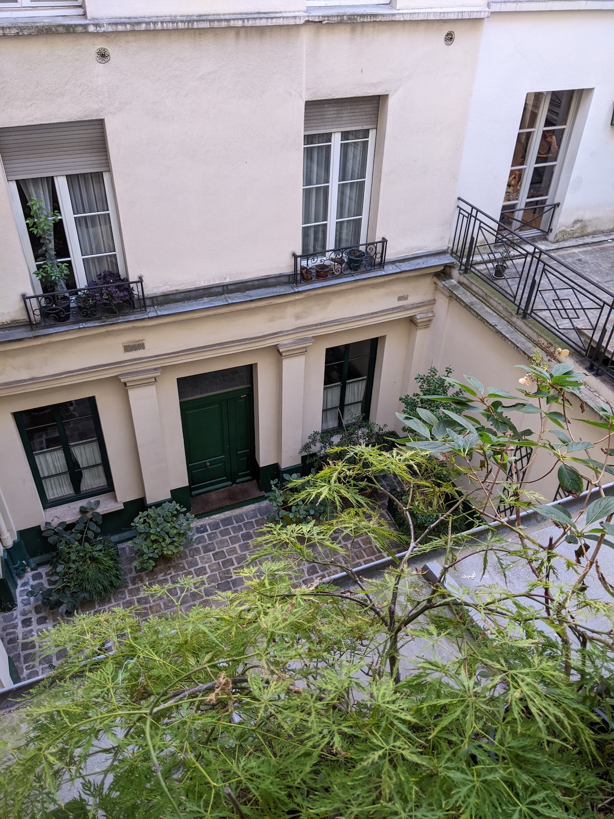 A view of a quiet, green courtyard is presented, featuring a doorway with a deep green door framed by light-colored walls. Plants adorn the space, enhancing the serene ambiance, while balconies above provide glimpses of additional greenery.