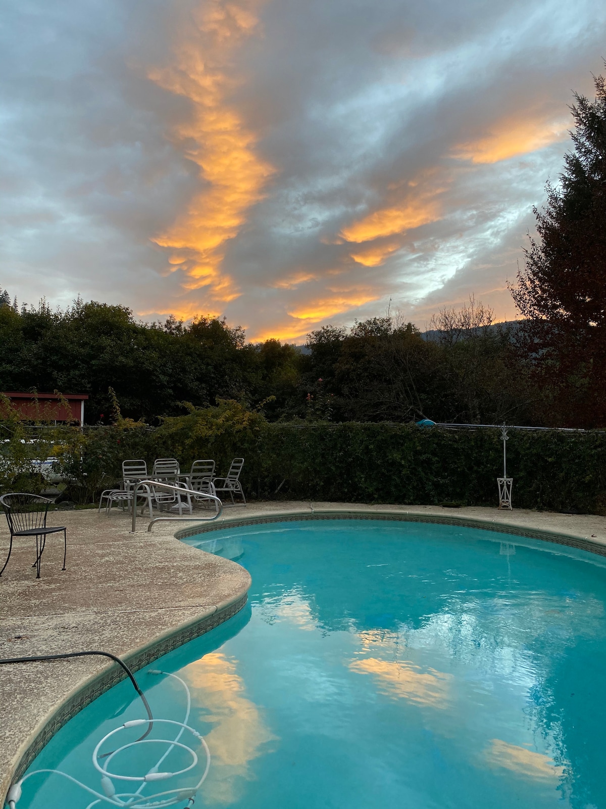 A glimmering saltwater pool reflects the colorful sky at sunset, featuring calming blues and warm orange hues. Surrounded by lush greenery, a spacious concrete deck hosts several lounge chairs positioned for relaxation.