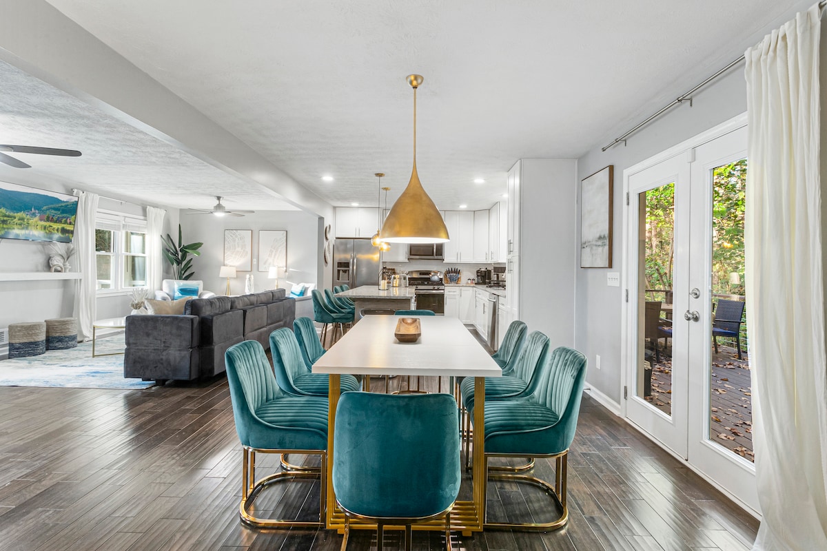 A dining area features a modern table surrounded by plush teal chairs. Natural light streams in through sliding glass doors, leading to an outdoor space. Pendant lighting hangs above the table, enhancing the open layout connecting to the stylish kitchen and living room.