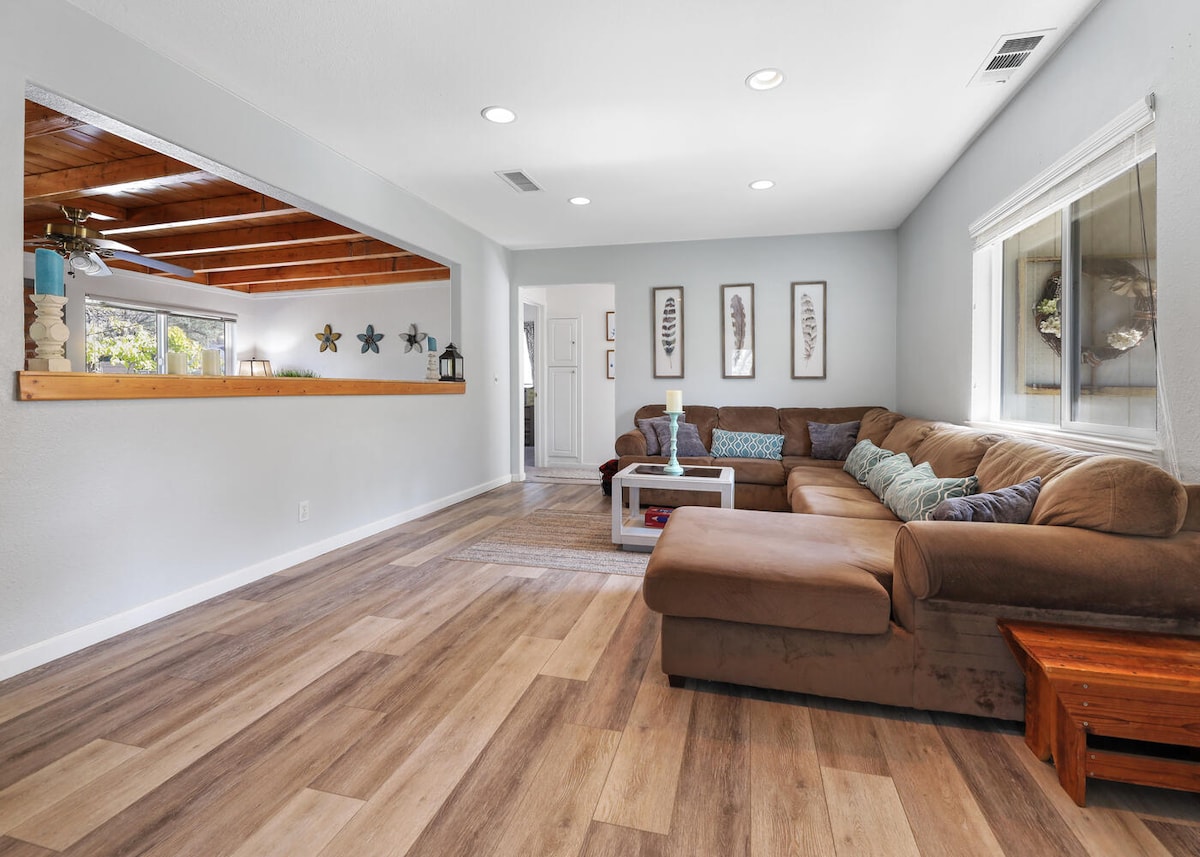 A spacious living area features a large L-shaped sectional sofa with colorful cushions. Light hardwood flooring complements the neutral walls. A white coffee table sits in front of the sofa, and decor can be seen on the wall, enhancing the cozy ambiance.