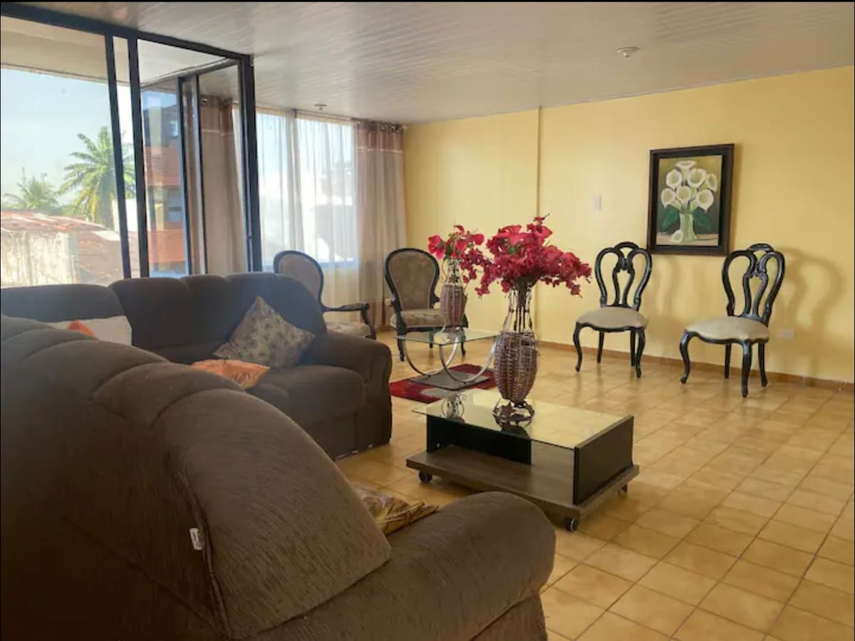 A spacious living area features a large, comfortable sofa and a coffee table adorned with decorative flowers. Several elegant chairs frame the space, complemented by large windows that allow natural light to fill the room, offering views of the surroundings.