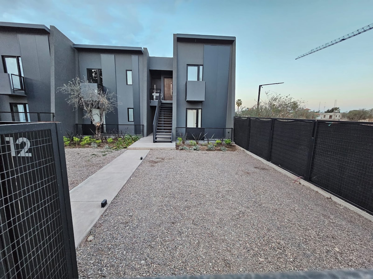 The modern building features a geometric design, with a gravel pathway leading to the entrance. Sparse landscaping includes low shrubs and small plants, while a fenced perimeter offers a sense of privacy. The structure has multiple levels, with stair access to upper units.