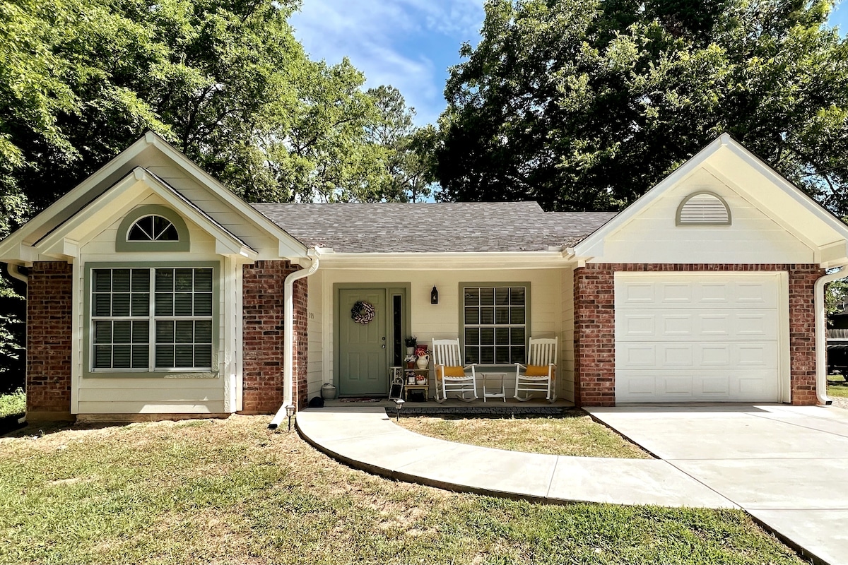 A charming cottage features a brick facade, complemented by light-colored trim and a welcoming front porch. Two rocking chairs are positioned beside the front door, while a garage is visible on the right. Lush greenery surrounds the property, enhancing the serene outdoor environment.