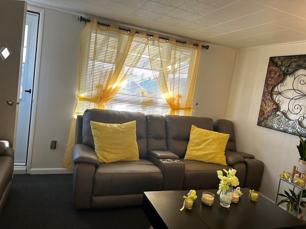 A cozy living room features a brown leather sofa adorned with yellow cushions. Natural light streams through sheer yellow curtains, illuminating the space. A dark coffee table is centered in front of the seating, decorated with candles and fresh flowers.