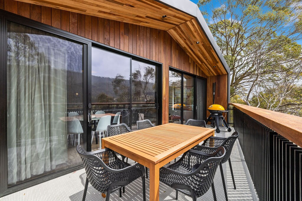 A spacious private balcony features a wooden dining table surrounded by six black mesh chairs. A barbecue grill is positioned nearby, with large glass doors leading into the living area. Natural light filters through, highlighting the wooden ceiling and outdoor greenery.