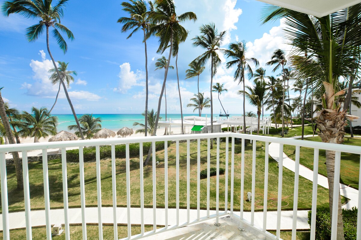 A spacious balcony overlooks a lush landscape dotted with palm trees swaying gently. The view extends to the sandy beach and turquoise ocean in the background, complemented by a clear blue sky filled with scattered clouds.