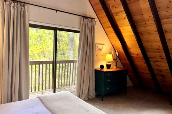 The primary bedroom features a king bed, antique dresser, and sliding glass doors that frame sweeping wooded views, creating a cozy retreat to unwind at the end of the day immersed in the peaceful wilderness surroundings.
