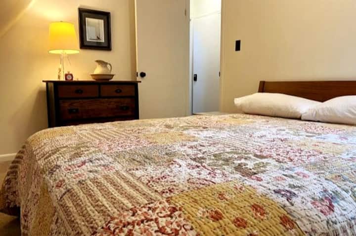 This charming upstairs bedroom provides a peaceful retreat with its queen bed, antique dresser, and rustic style embraced by the natural beauty of the surrounding woods.