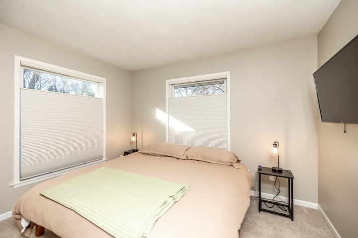 Მთავარი იატაკის საძინებელი დიდი საწოლით, მყუდრო თეთრეულით, რბილი განათებით და კედელზე მიმაგრებული ტელევიზორით, რომელიც იდეალურია კომფორტული დასვენებისთვის.
