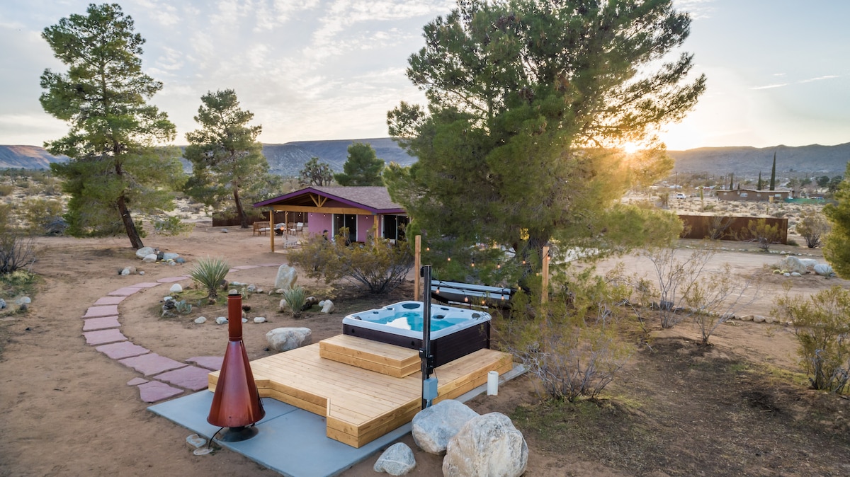 An outdoor area showcases a cowboy tub soaking pool adjacent to a wooden deck and a propane fireplace. A pathway lined with stones leads through the desert landscape, framed by desert vegetation and trees, while the sunset casts a warm glow over the surroundings.