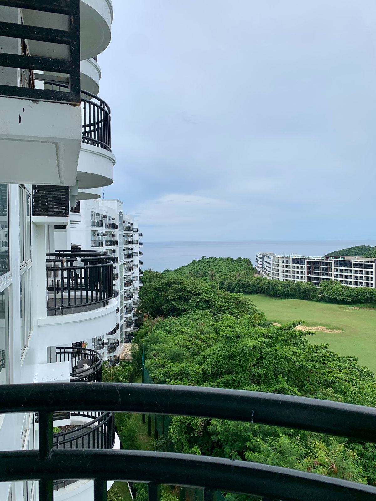 A view from a private balcony overlooking lush greenery and the azure ocean. The undulating coastline and nearby golf course are visible, with modern condominium buildings framing the scene. The sky is partially cloudy, creating a serene atmosphere.