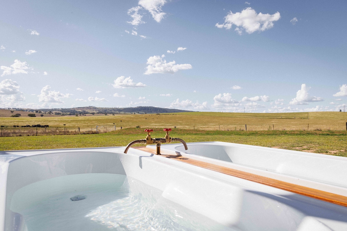An outdoor hot tub is presented with a wooden plank alongside it, offering a view of expansive green pastures and rolling hills under a partly cloudy sky. The serene landscape complements the tranquil bath setting.