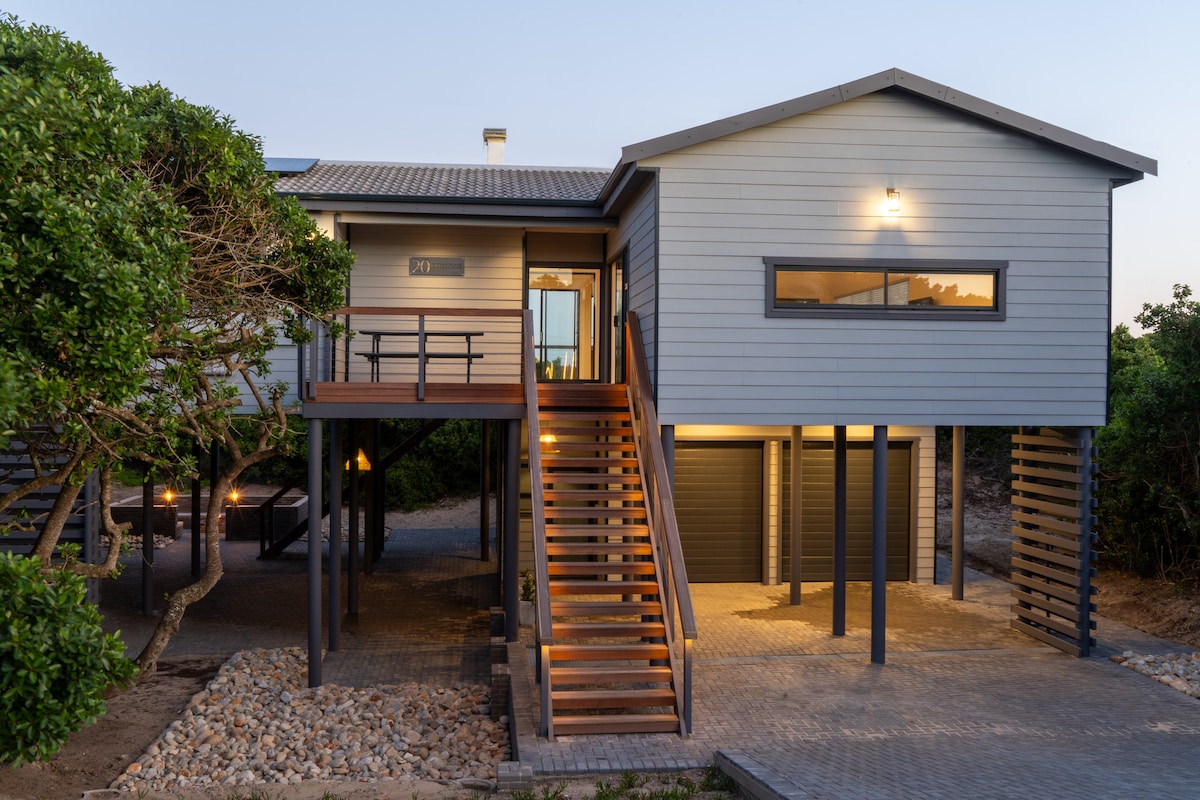 The exterior of a modern, elevated house features a wooden staircase leading to a spacious deck. Flanking the entrance are large windows, allowing natural light to fill the interior. The surrounding landscape includes greenery and a stone pathway, providing a welcoming approach.