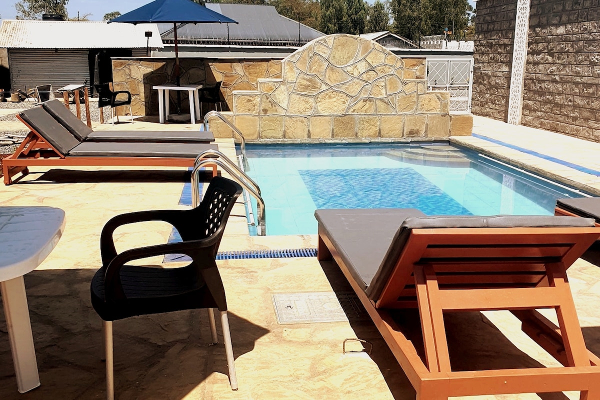 A refreshing swimming pool is positioned in a sunlit area, surrounded by comfortable sunbeds. The stone wall adds a natural touch, while an umbrella provides shade nearby for relaxation. A small table is visible in the background, offering a space for refreshments.