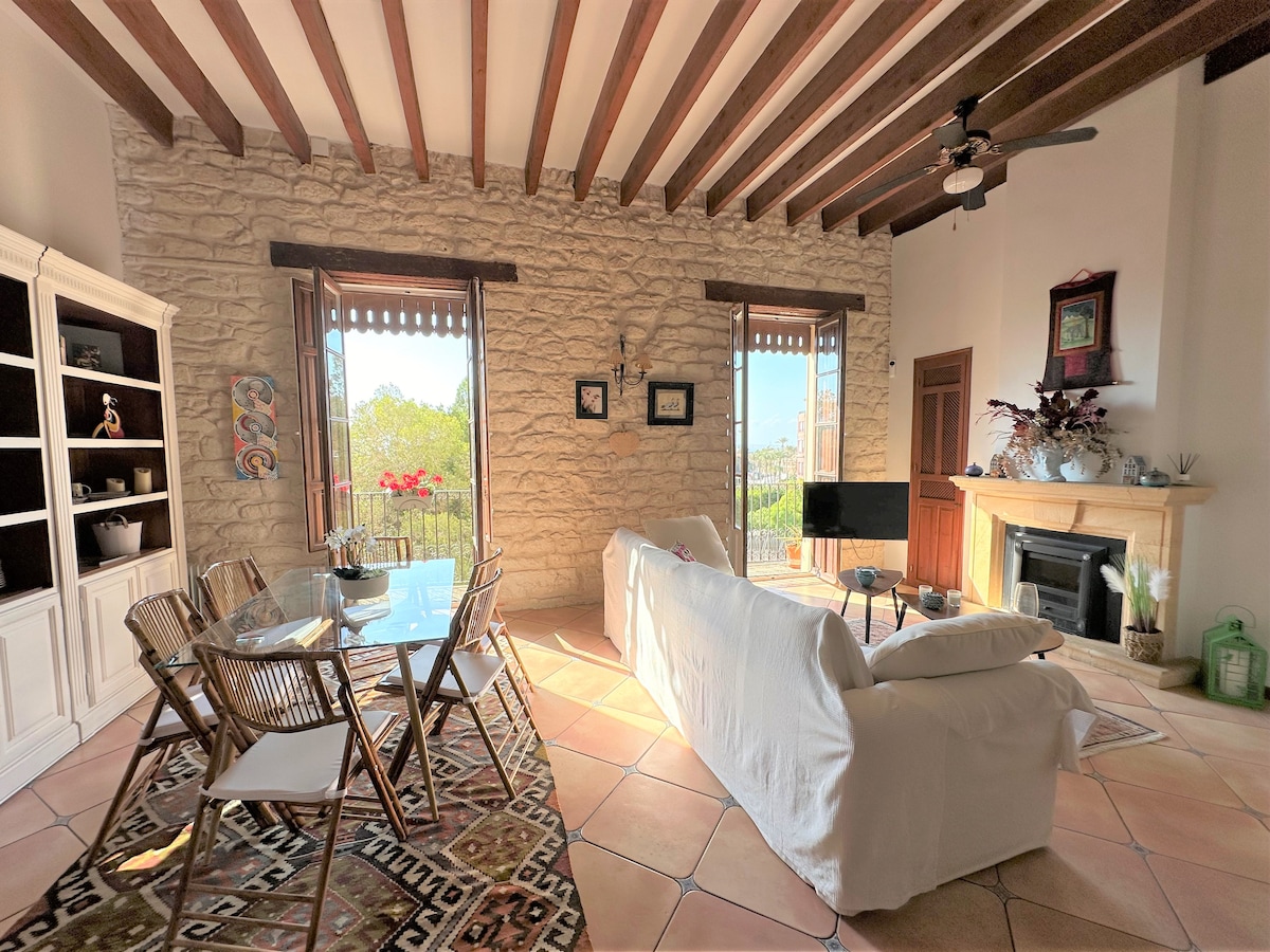 A spacious living area features exposed wooden beams and stone walls, complemented by large windows. A glass dining table and chairs are positioned in a sunlit corner, while a comfortable sofa faces a fireplace, creating a cozy gathering space.