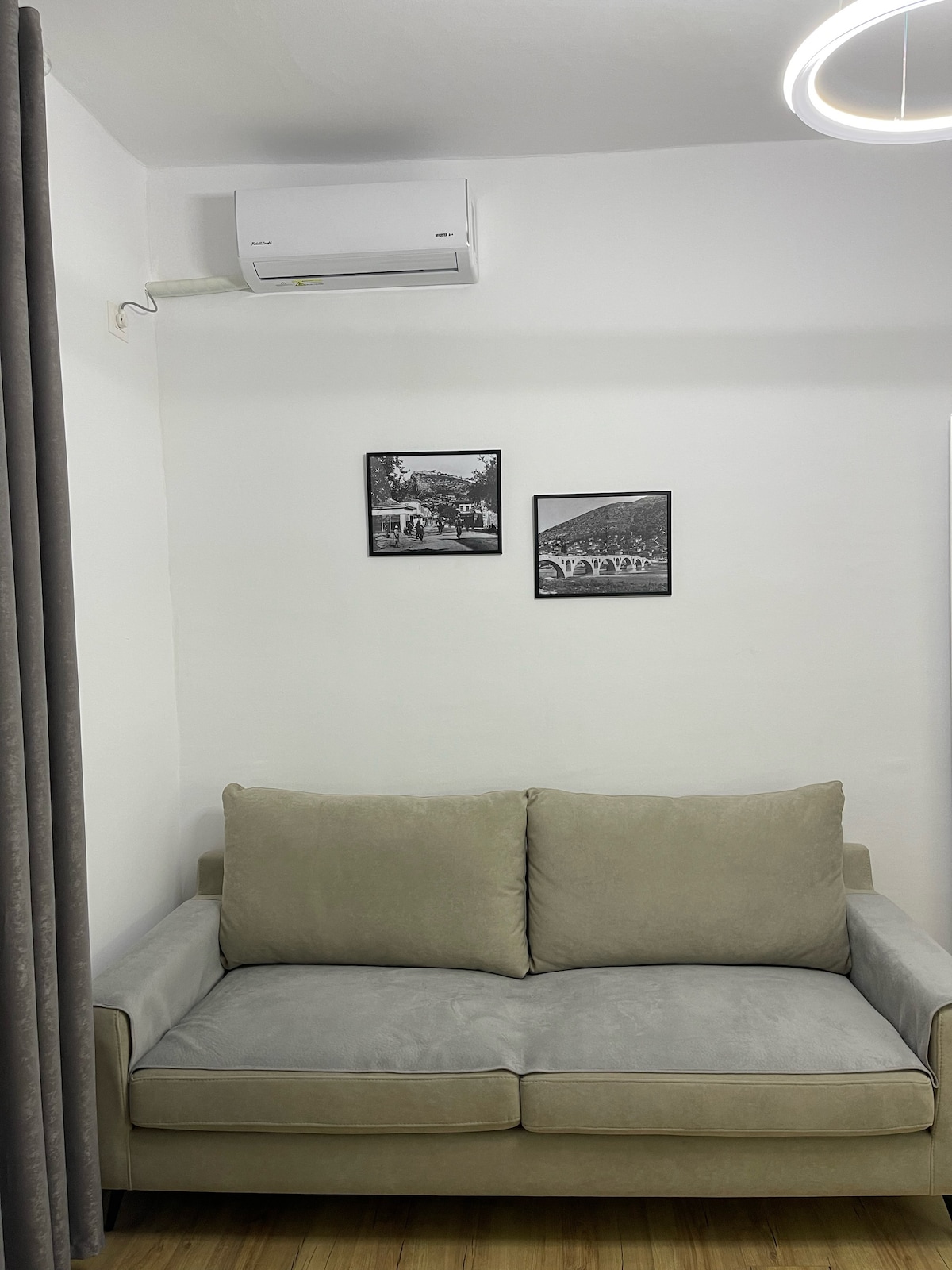 A spacious sofa is positioned against a white wall, accompanied by two framed black and white photographs above it. An air conditioning unit is mounted on the wall, ensuring a comfortable environment.