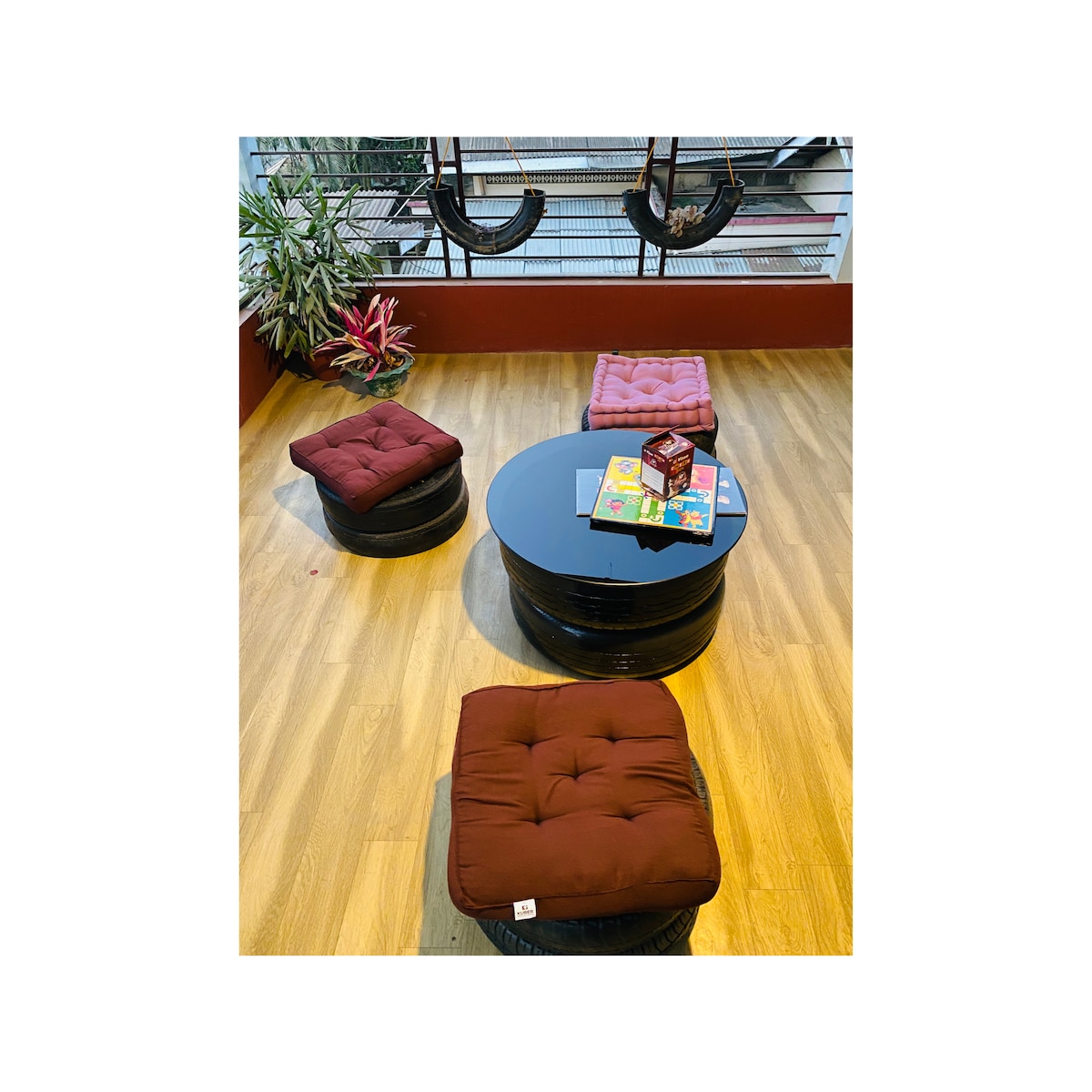 A cozy seating area features a round black coffee table surrounded by four cushioned stools in warm tones. The space is brightened by natural light streaming through large windows, with plants adding a touch of greenery. Two hanging chairs offer additional seating options.