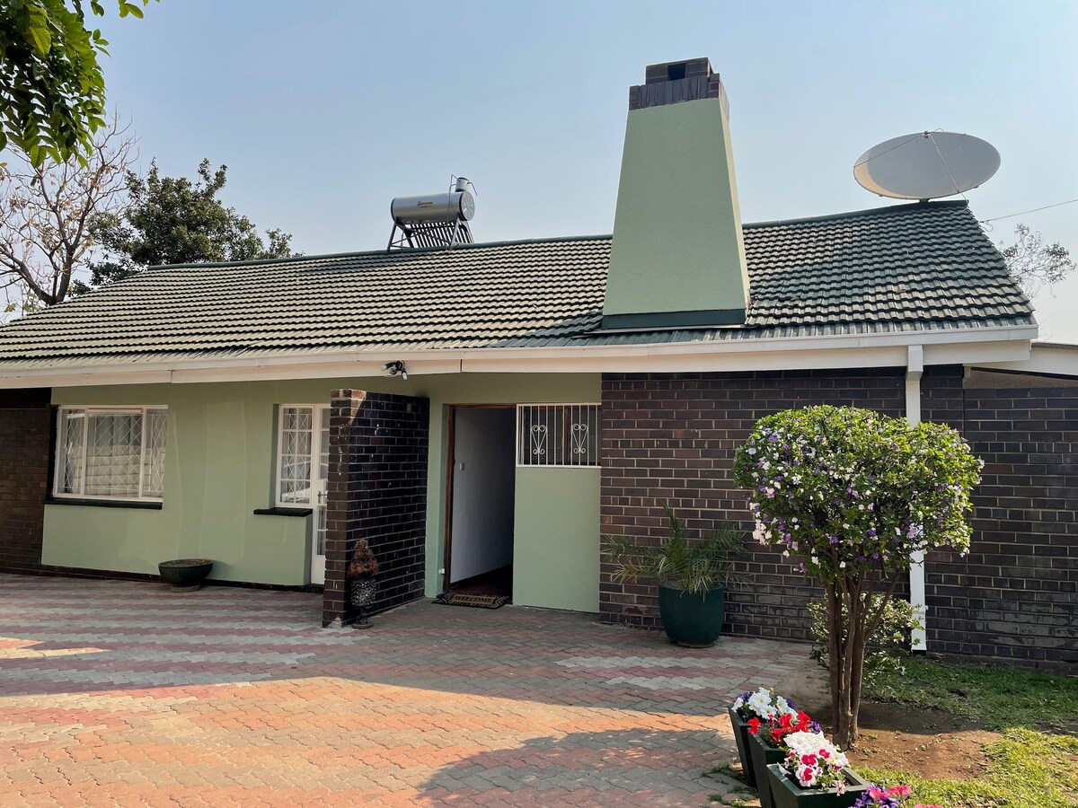 The exterior of a single-story home is shown, featuring a well-maintained brick facade with light green accents. A traditional chimney rises from the roof, beside a satellite dish. A flowerbed with blooming plants complements the entrance, while a paved walkway leads to the front door.