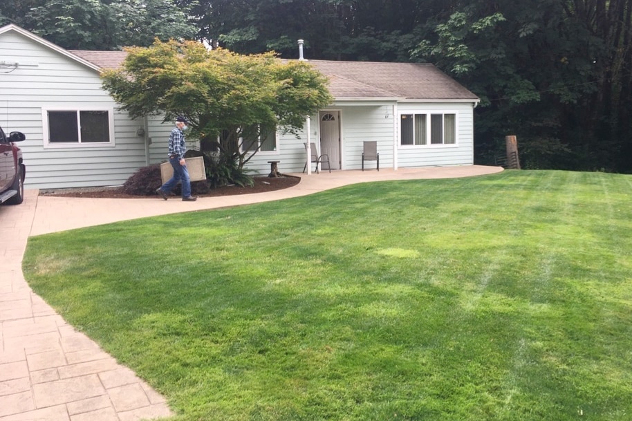 A single-story home is set in a lush green landscape, with a spacious lawn and patio area. Two chairs are positioned outside the entrance. A tree provides partial shade, and the exterior features light-colored siding with multiple windows, reflecting a serene environment.