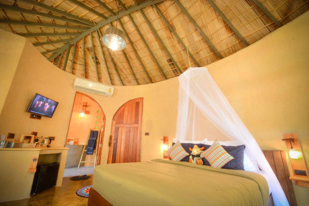 A round guest room features a thatched ceiling and earthy-toned walls. A spacious bed is dressed with patterned pillows and a lightweight throw. A flat-screen TV is mounted on the wall, alongside an entryway leading to a private bathroom with modern amenities.
