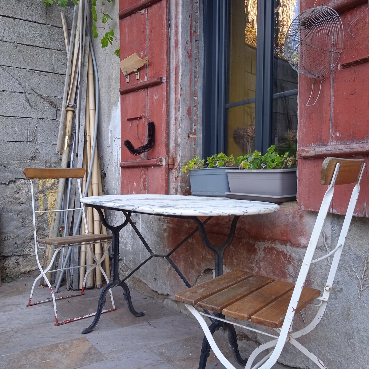 Two wooden chairs are positioned around an antique round table on a stone patio. The rustic wall, painted in red and gray, provides a backdrop, while potted plants add a touch of greenery. Garden tools can be seen stacked nearby.