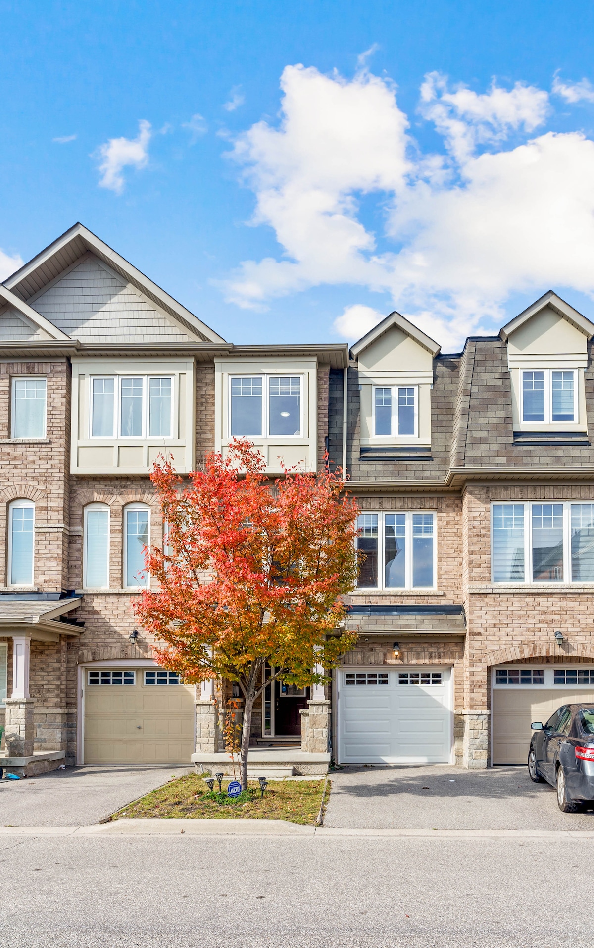 The townhouse exterior is showcased, featuring a blend of brick and siding materials. A vibrant red tree stands at the entrance, adding a pop of color to the scene. Large windows allow natural light to illuminate the home, and vehicle parking is visible in the driveway.