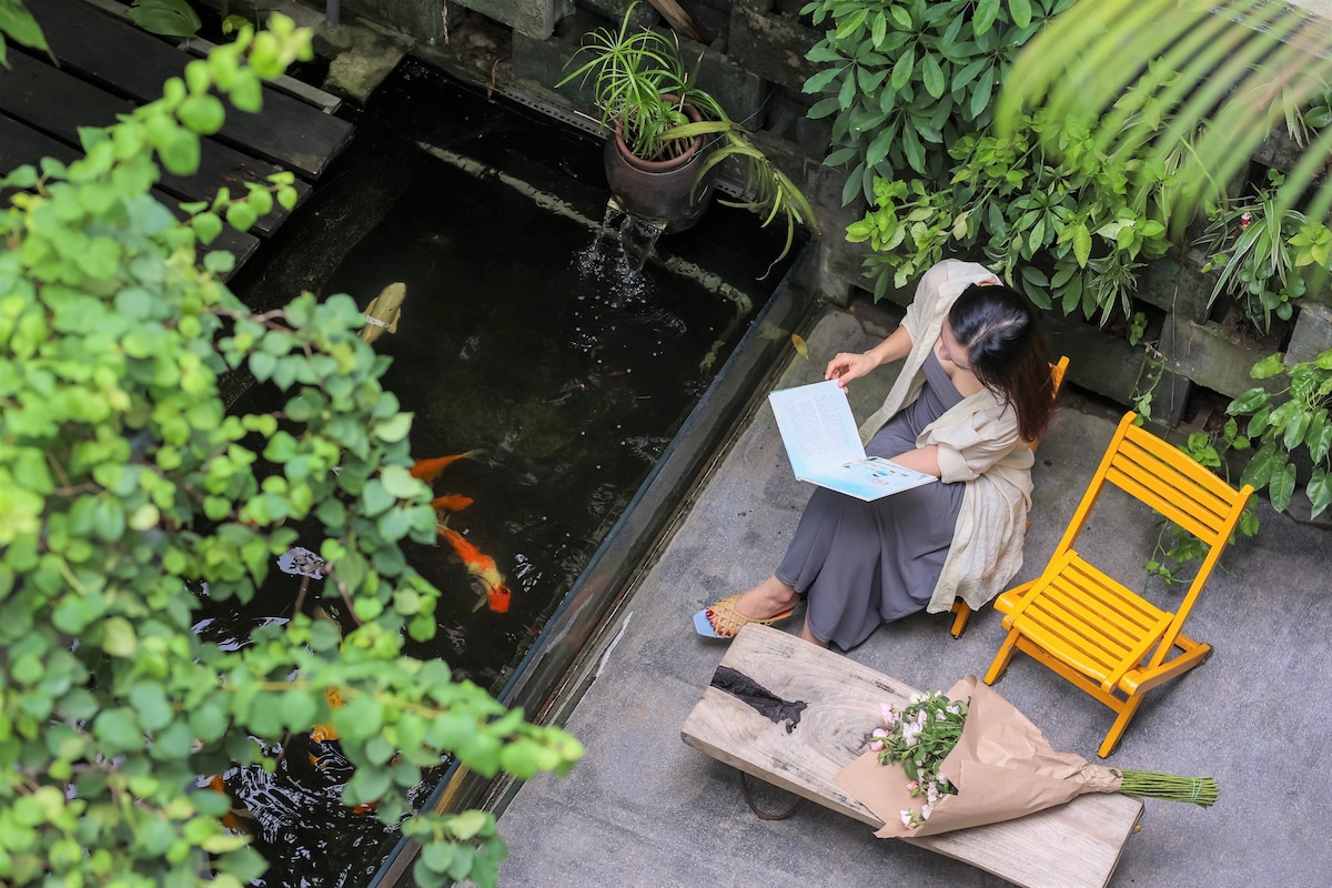 A tranquil outdoor space features a small Koi pond surrounded by lush greenery. A person sits on a bright yellow chair, engaged in reading a book. Nearby, a bouquet of flowers rests on the surface, adding a natural touch to the serene environment.