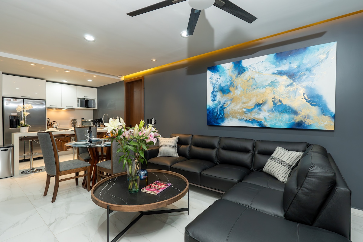 A modern living area features a large black sectional sofa and a round marble coffee table. A colorful abstract painting adorns the grey wall, while a dining table for four sits near the open kitchen. Natural light illuminates the space, creating a welcoming ambiance.