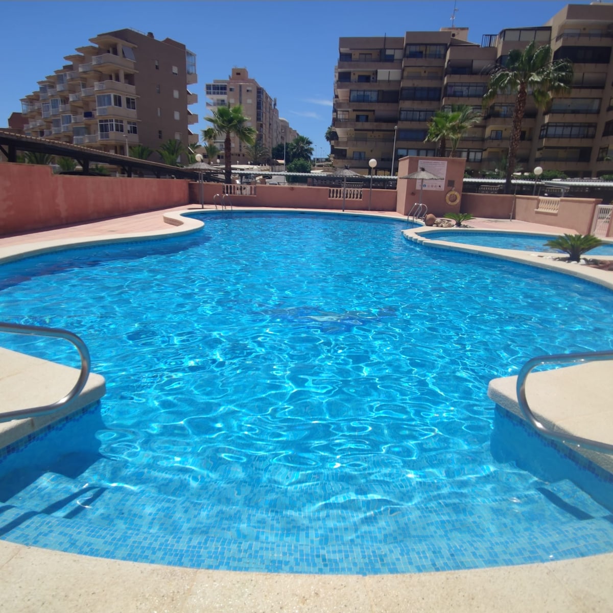 A clear blue swimming pool is framed by a light-colored deck, with steps leading into the water. Surrounding palm trees provide shade, while the backdrop features several multi-story buildings under a clear sky. Natural light enhances the inviting appearance of the pool area.